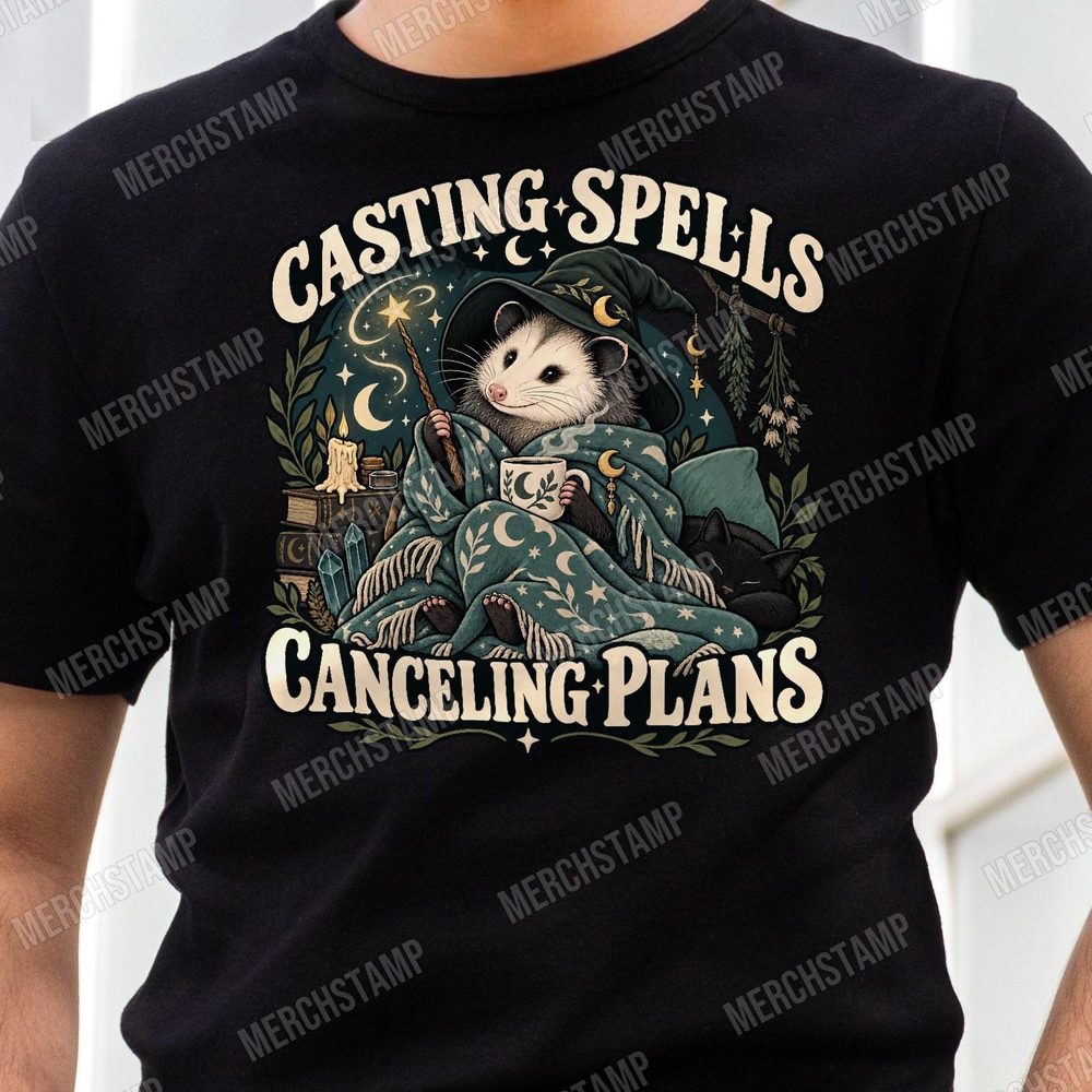 Casting Spells Canceling Plans PNG design shown on a black t-shirt, a sleepy opossum in a wizard hat wrapped in a starry blanket with a wand and a mug, between the words Casting Spells Canceling Plans