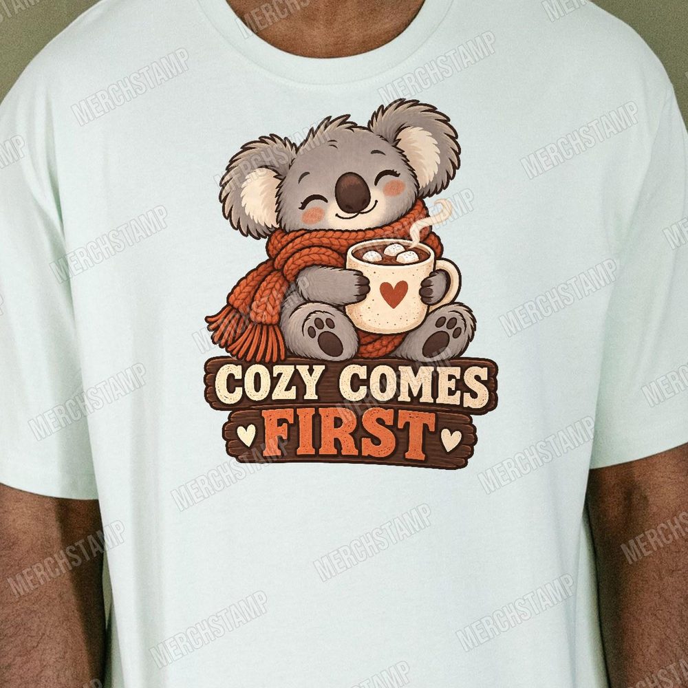 Cozy Comes First PNG design shown on a pale mint t-shirt, a sleepy koala in an orange scarf hugging a mug of cocoa above the words Cozy Comes First